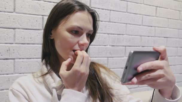 Piękna Brunetka kobieta oglądanie straszne seria seryjnego w słuchawkach na telefon komórkowy na tle białej ściany Cegła. — Wideo stockowe