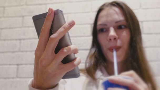 Mosolygó gyönyörű nő iszik egy italt egy cső, és néz video-ra mozgatható telefon-ban kávézó. — Stock videók