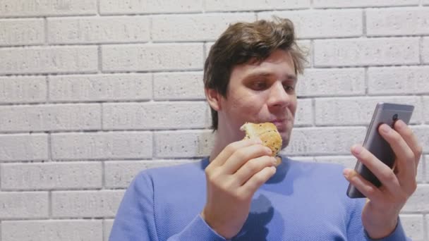 El hombre está comiendo una hamburguesa en la cafetería y viendo un video en el teléfono móvil . — Vídeo de stock