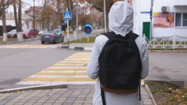 Kvinnan i vit jacka med ryggsäck kommer att korsa vägen vid ett övergångsställe i staden. — Stockvideo