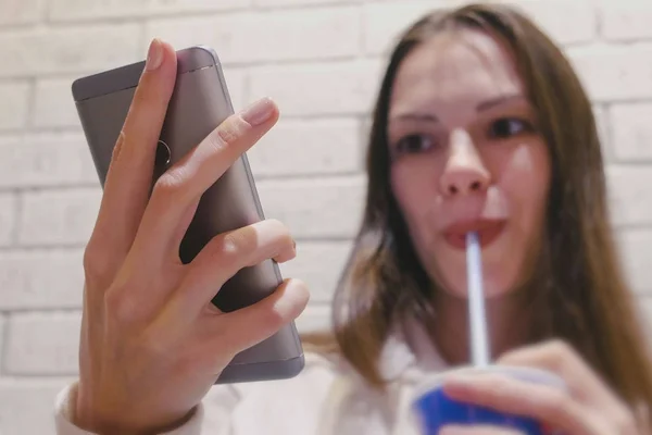 Sonriendo hermosa mujer está bebiendo una bebida de un tubo y viendo vídeo en el teléfono móvil en la cafetería . — Foto de Stock