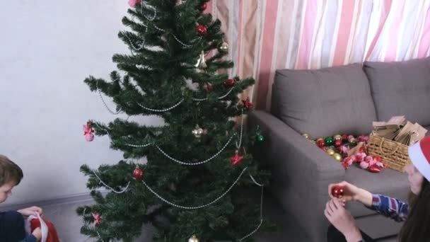 Μαμά και γιος στο Χριστούγεννα καπέλα διακοσμήσετε το χριστουγεννιάτικο δέντρο με χάντρες και μπάλες. — Αρχείο Βίντεο