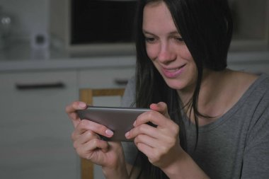 Beautiful young woman stealthily playing games on mobile phone in the kitchen at night. clipart