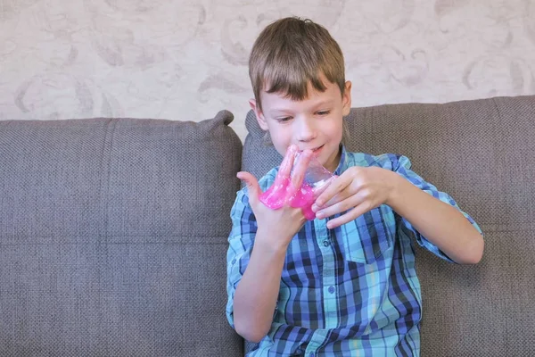 Happy boy is putting a pink slime on his hand sitting on the coach and playing. — Stock Photo, Image
