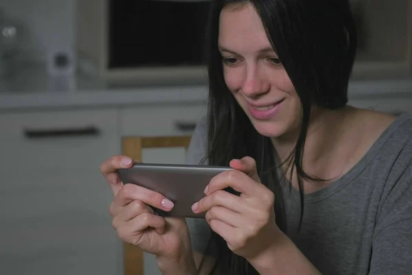 Hermosa mujer joven sigilosamente jugando juegos en el teléfono móvil en la cocina por la noche . — Foto de Stock