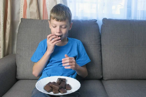 Tiener jongen is het eten van chocolade snoepjes zittend op de Bank. Concept van ongezond eten. — Stockfoto