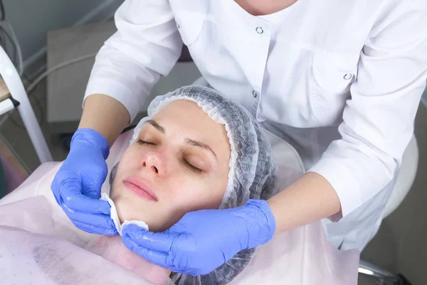 Cosmetologo pulisce il viso di una giovane donna con batuffoli di cotone. Mani dell'estetista in guanti e un primo piano di faccia. Trattamenti di bellezza per il viso . — Foto Stock