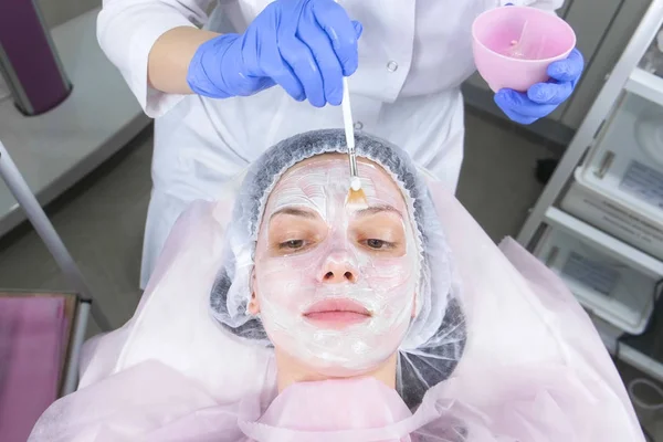 Kosmetolog sætter en hvid maske på kvindens ansigt med en børste. Hænderne på en kosmetolog i blå gummihandsker. Ansigtsbehandlinger . - Stock-foto