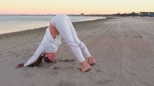 Молодая женщина занимается йогой Adho mukha shvanasana на песчаном пляже на рассвете осенью . — стоковое видео