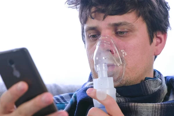 Vernebler und Inhalator für die Behandlung verwenden. Kranker Mann inhaliert durch Inhalationsmaske und schaut auf Handy auf dem Sofa sitzend. Gesicht aus nächster Nähe. — Stockfoto