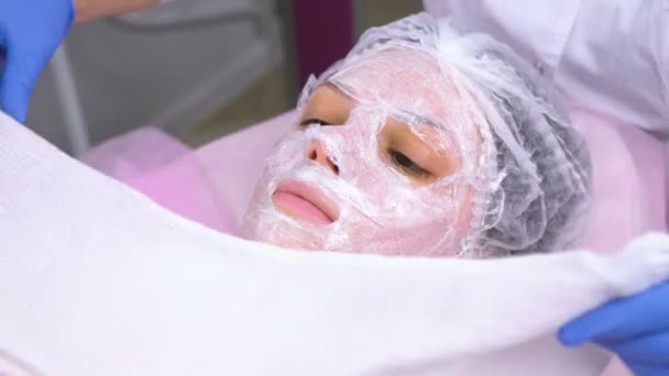 Kosmetyczka ściąga womans twarz ręcznikiem. Kosmetolog stawia folię na szczycie nawilżająca maseczka na twarz womans. — Wideo stockowe