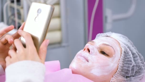 Woman reads something in a mobile phone lying on the procedure in the cosmetoloists office with a mask on her face. — Stock Video