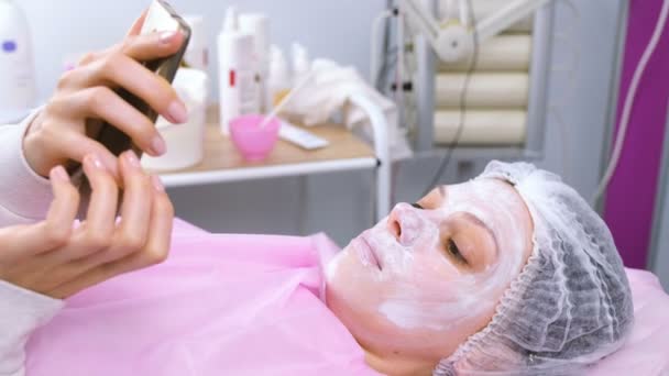 Kvinnan läser något i en mobiltelefon som liggande på förfarandet i cosmetoloists kontoret med en mask på ansiktet. — Stockvideo