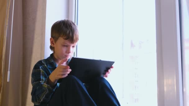Seroisly menino está lendo livro em tablet sentado no peitoril da janela . — Vídeo de Stock
