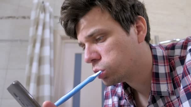 Сумасшедший молодой человек чистит зубы и смотрит на телефон в ванной . — стоковое видео