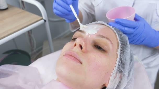 Beautician menaruh topeng putih di wajah wanita dengan kuas. Tangan seorang ahli kosmetologi sarung tangan karet biru. Perawatan wajah . — Stok Video