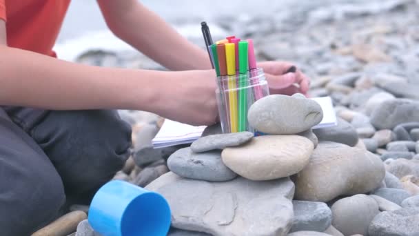 Close-up boys manos está pintando con marcadores mientras está sentado en una playa de piedra en la orilla del mar . — Vídeo de stock
