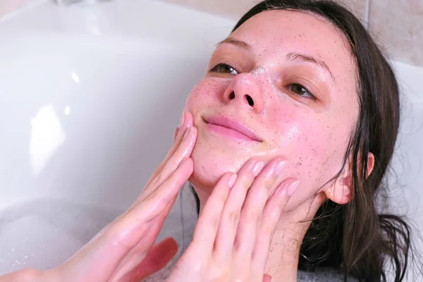 Hermosa mujer morena pone un exfoliante en la cara acostada en el baño. Primer plano de la cara . — Foto de Stock