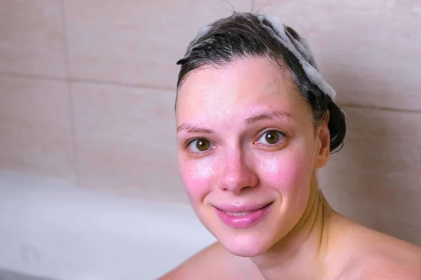 Vrouw is wassen haar hoofd en haren met shampoo in de badkamer. Kijken in de camera. — Stockfoto