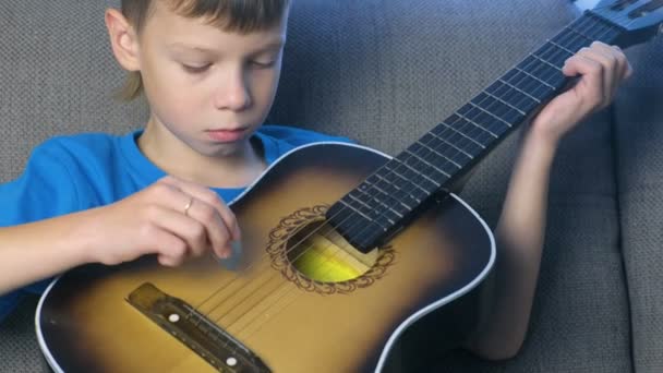 Boy adalah bermain gitar duduk di sofa. Konsep belajar untuk memainkan alat musik . — Stok Video