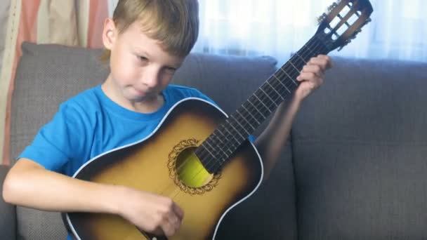 Pojke lär sig att spela gitarr som sitter i soffan. Begreppet att lära sig spela ett musikinstrument. — Stockvideo