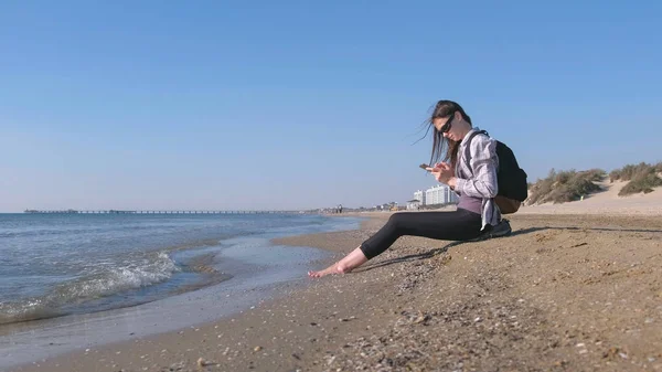 Chica viajera con una mochila sentada en una playa de arena y escribiendo un mensaje en el teléfono móvil . —  Fotos de Stock