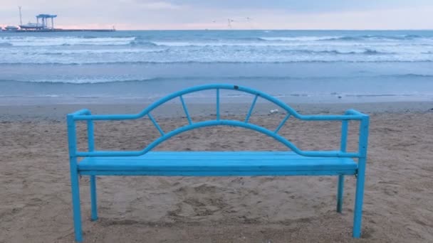 Kumlu sahilde günbatımı mavi bankta. Deniz kenarında rahatlamak için uygun altyapı. Dalga deniz. — Stok video