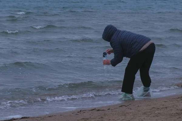 Femme âgée voyageur gagne une bouteille d'eau de mer debout sur une plage de sable. Automne au bord de la mer. Souvenir de mer . — Photo