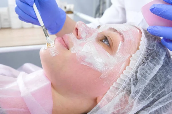 La esteticista pone una máscara blanca en la cara de las mujeres con un cepillo. Vista lateral. Manos de un cosmetólogo con guantes de goma azul. Tratamientos faciales . — Foto de Stock