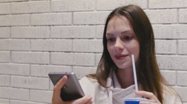 Gülümseyen güzel kadın bir tüp sizden bir içecek içme ve cep telefonu kafede video izlerken.