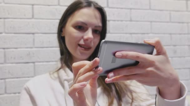 Mooie brunette vrouw kijken naar een seriële in koptelefoon op mobiele telefoon op een bakstenen muur achtergrond wit. — Stockvideo