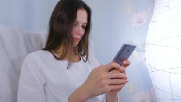 Joyeux jeune femme brune tape un message sur un téléphone portable à la maison . — Video