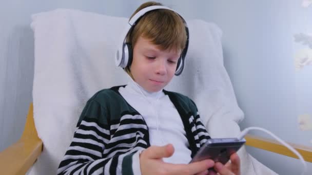 Pojken är musik i hörlurar i smartphone som sitter i fåtöljen hemma. — Stockvideo