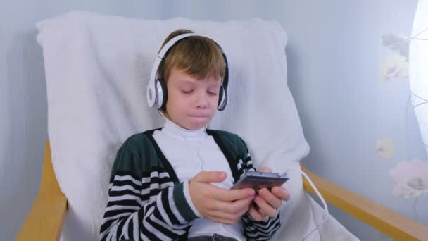 Boy está escuchando música en los auriculares en el teléfono inteligente y cantando sentado en el sillón en casa . — Vídeos de Stock