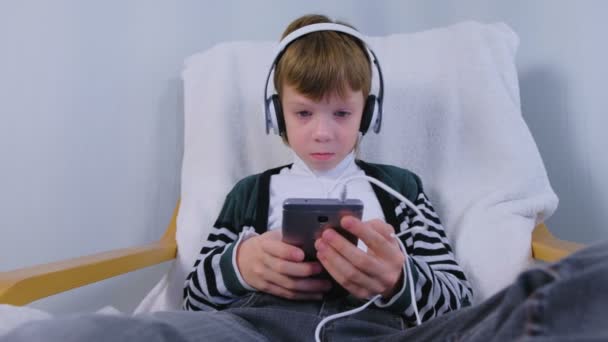 Boy está escuchando música en los auriculares en el teléfono inteligente sentado en el sillón en casa . — Vídeo de stock