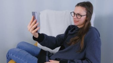Mutlu genç esmer kadın gözlük koltukta oturan bir cep telefonunda görüntülü sohbet konuşmak.