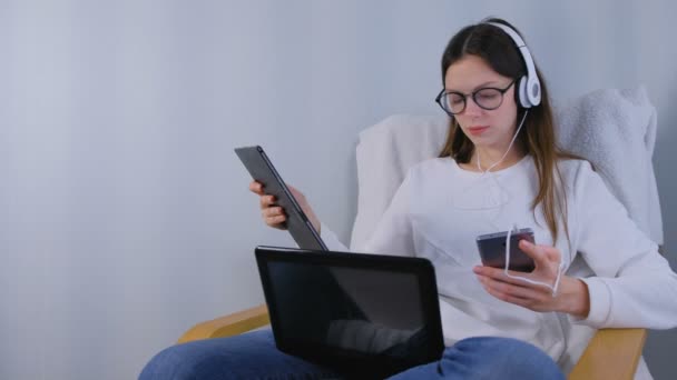 Жінка в окулярах і навушниках працює на ноутбуці, сидячи в кріслі. Мобільний телефон і планшет в руках . — стокове відео
