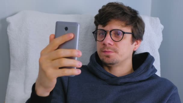 Mann mit Brille spricht Videochat auf Smartphone im Sessel. — Stockvideo