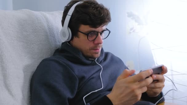 Чоловік в окулярах і навушниках грає в гру в смартфоні. Гамер розслабляється . — стокове відео