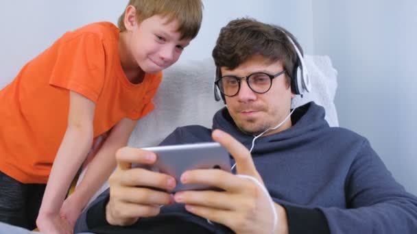 Man in glazen en hoofdtelefoon spelen van een spel in smartphone met zijn zoon. Gamer ontspannen. — Stockvideo