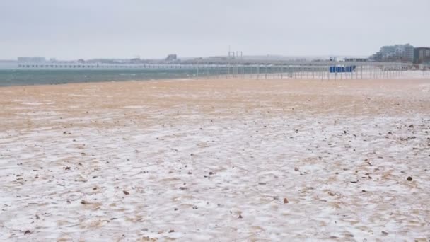 Snow in winter on the sea sand beach with coastal town on background. — Stock Video