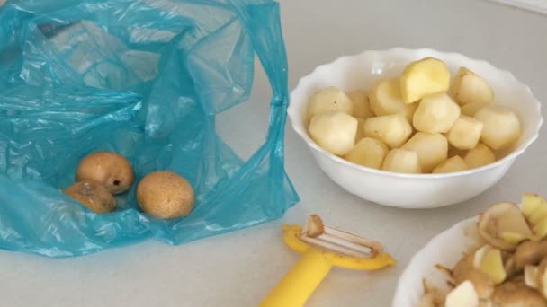 Cartofi mici în farfurie, coajă și peeler pe masa de bucătărie. Cartofi nedecojiți pe punga albastră de plastic . — Videoclip de stoc