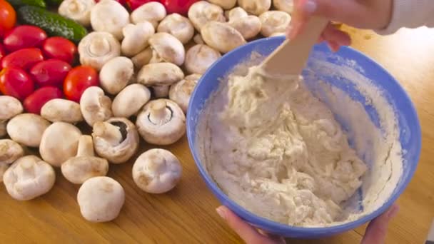 Mani delle donne sta impastando la pasta di lievito con una spatola di legno in ciotola blu sul tavolo con verdure . — Video Stock