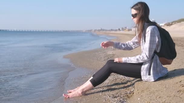 Utas a egy hátizsák, ül egy homokos strand és a tengerre néz. Fiatal lány a strandon. — Stock videók