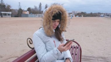 Sahilde bir bankta oturmuş ve üzgün video cep telefonu seyir ceket aşağı beyaz bir kadın.