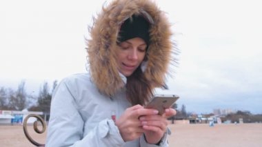 Sahilde bir bankta oturmuş ve cep telefonunda bir mesaj yazarak ceket aşağı beyaz bir kadın.