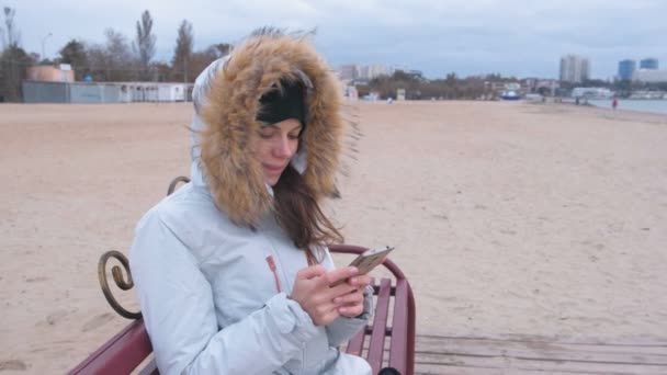 Vrouw in een witte donsjack zittend op een bankje op het strand en kijken naar mobiele telefoon. — Stockvideo