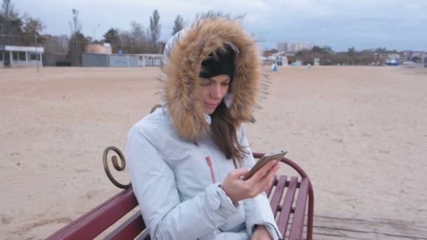 ダウン ジャケット浜辺のベンチに座って、携帯電話を悲しいビデオを見て白衣の女. — ストック動画