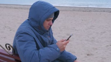 Adam blogger bir mavi Kum Plajı üzerinde bir bankta oturmuş ve cep telefonu sosyal ortamda yazı ceket aşağı.