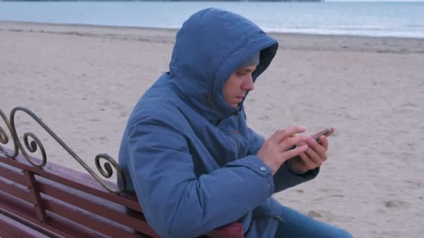 Hombre en una chaqueta azul sentado en un banco en la playa de arena, llamando al teléfono móvil y esperando respuesta . — Vídeo de stock